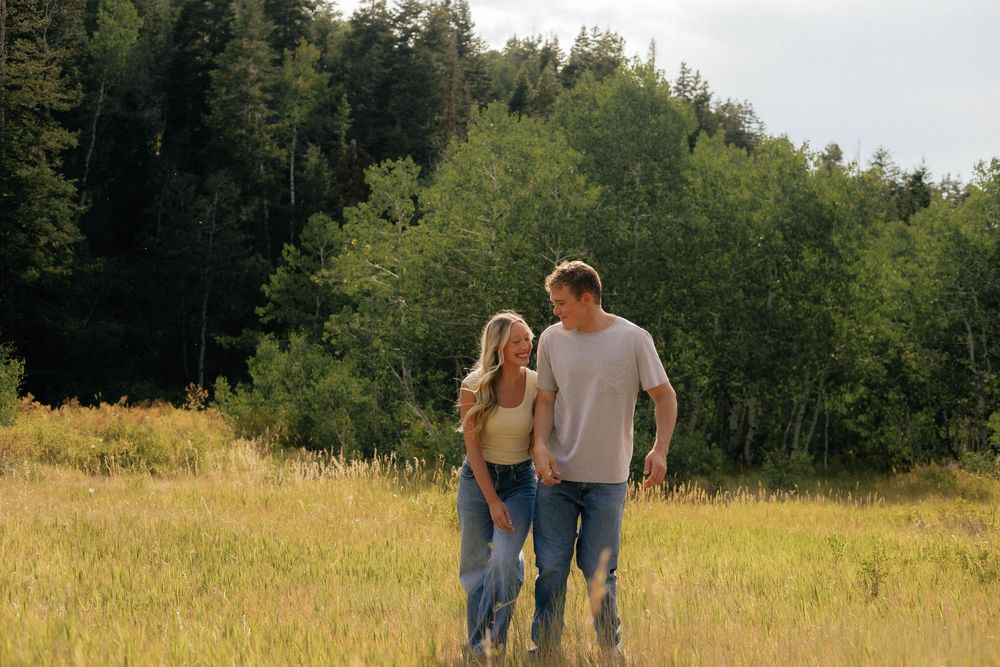 A couple walks hand in hand through a sunny meadow with lush green trees in the background.