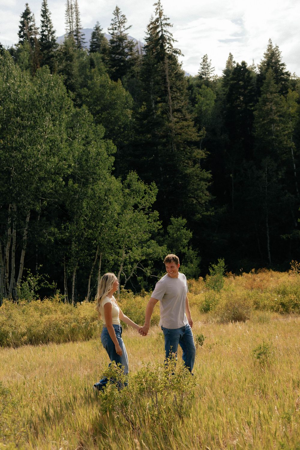 Young couple holding hands while walking through a golden meadow with evergreen forest backdrop.
