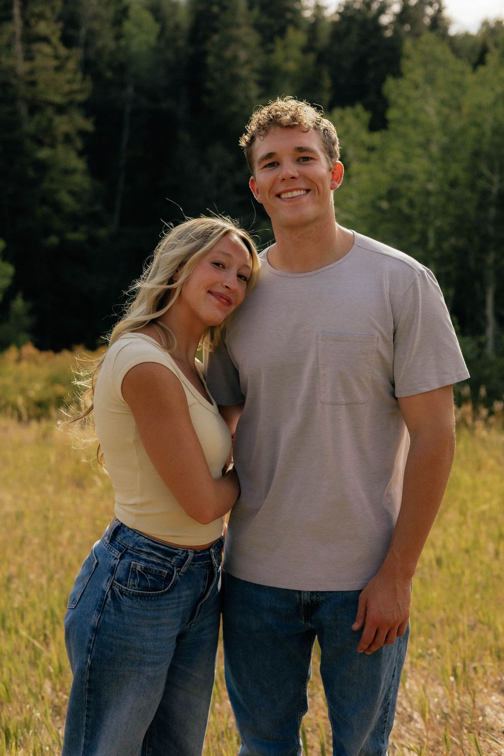 Smiling young couple posing together in a sunlit mountain meadow surrounded by tall pine trees.