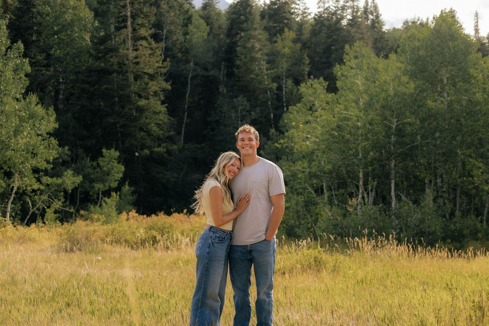 Couple smiling together in a sunny meadow surrounded by lush green trees and golden grass.