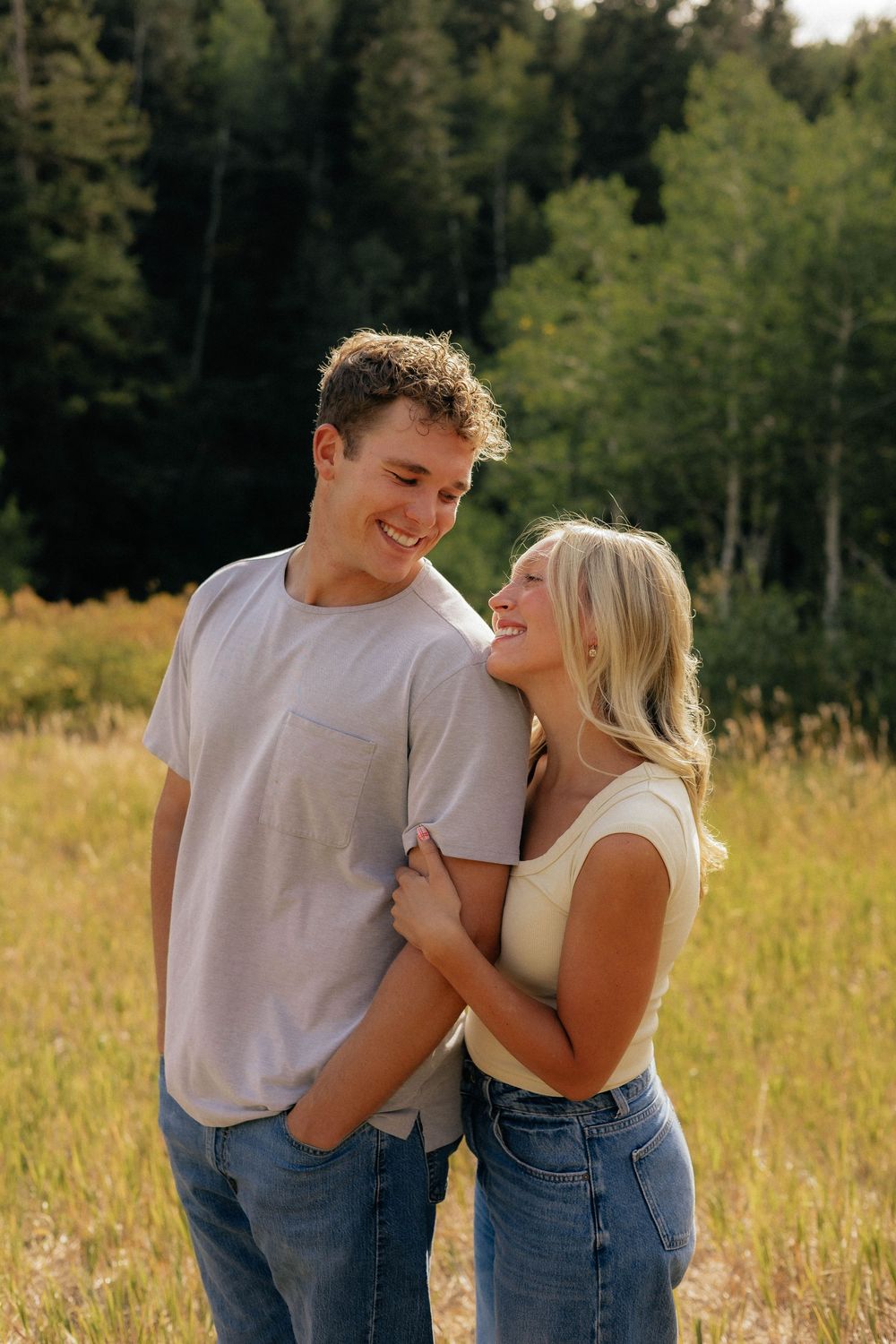 Happy young couple laughing and embracing in a golden field with a lush green forest behind them.