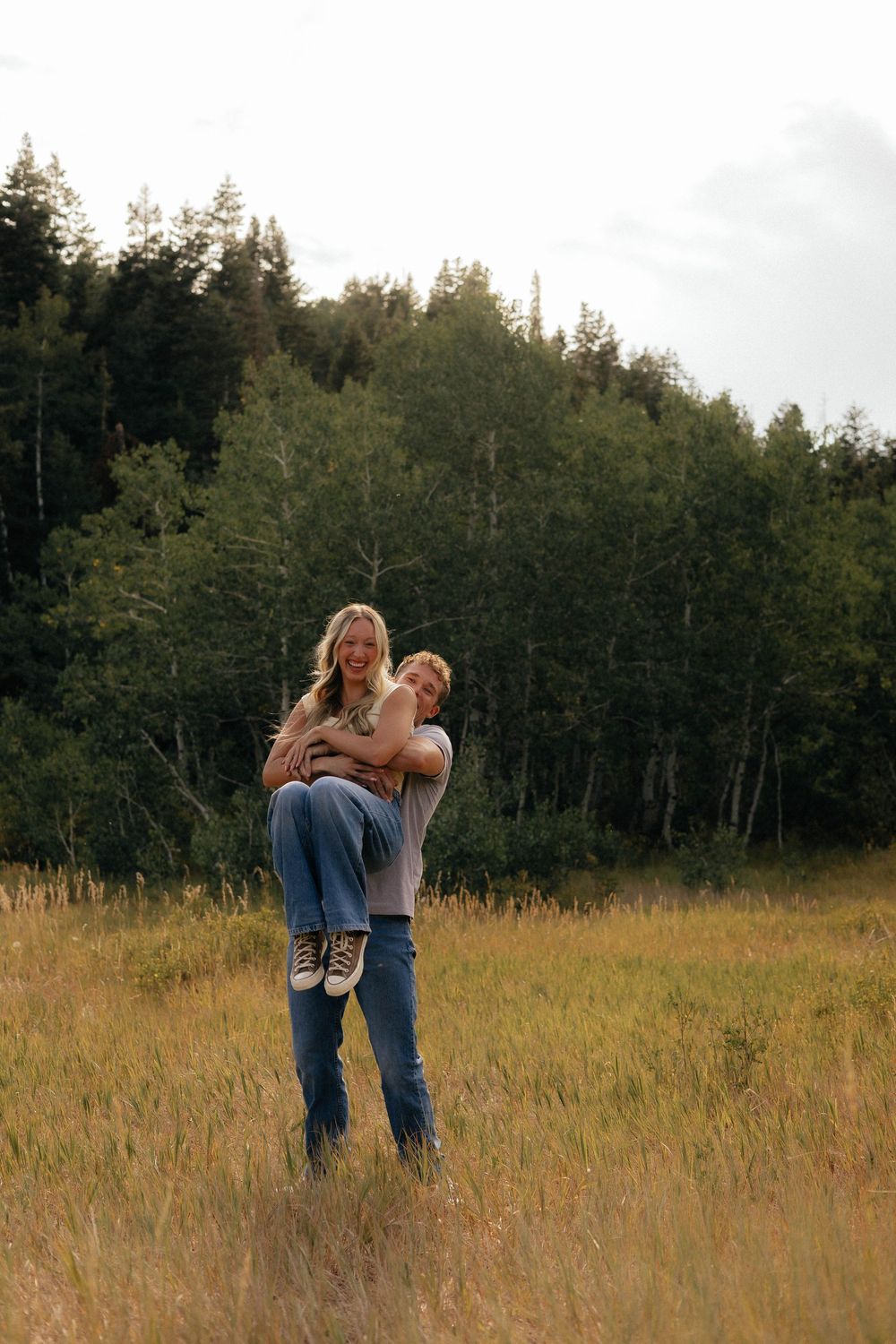 A man lifts a smiling woman in a golden meadow with lush green trees in the background.