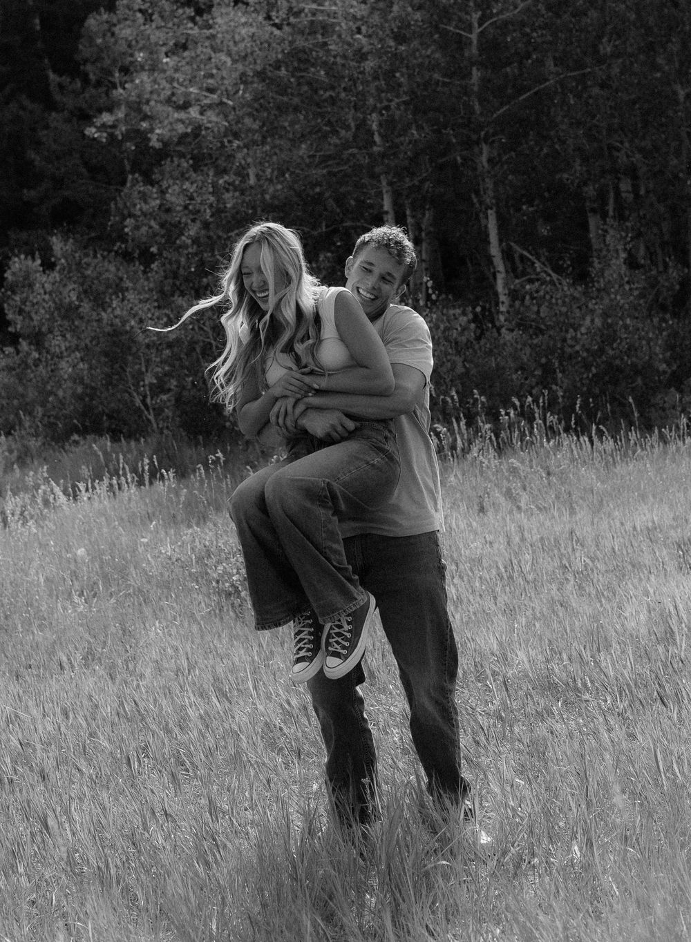 Black and white photo of a couple laughing in a grassy field, man lifting woman playfully.