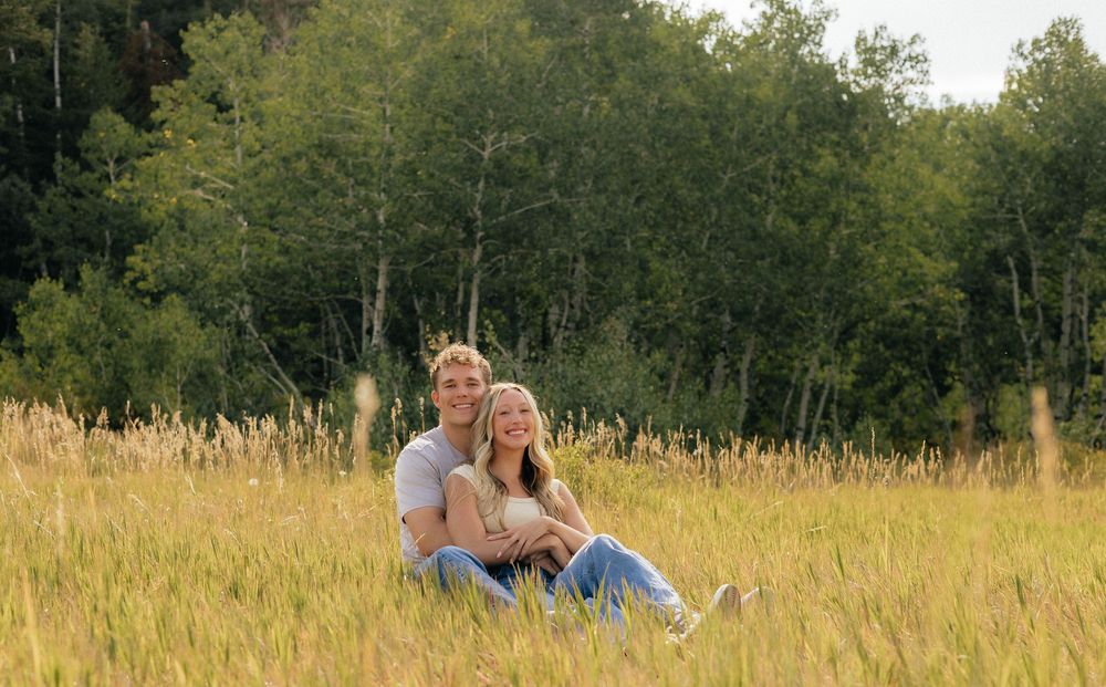 Couple sitting together in a golden meadow field with lush green trees in the background on a sunny day.