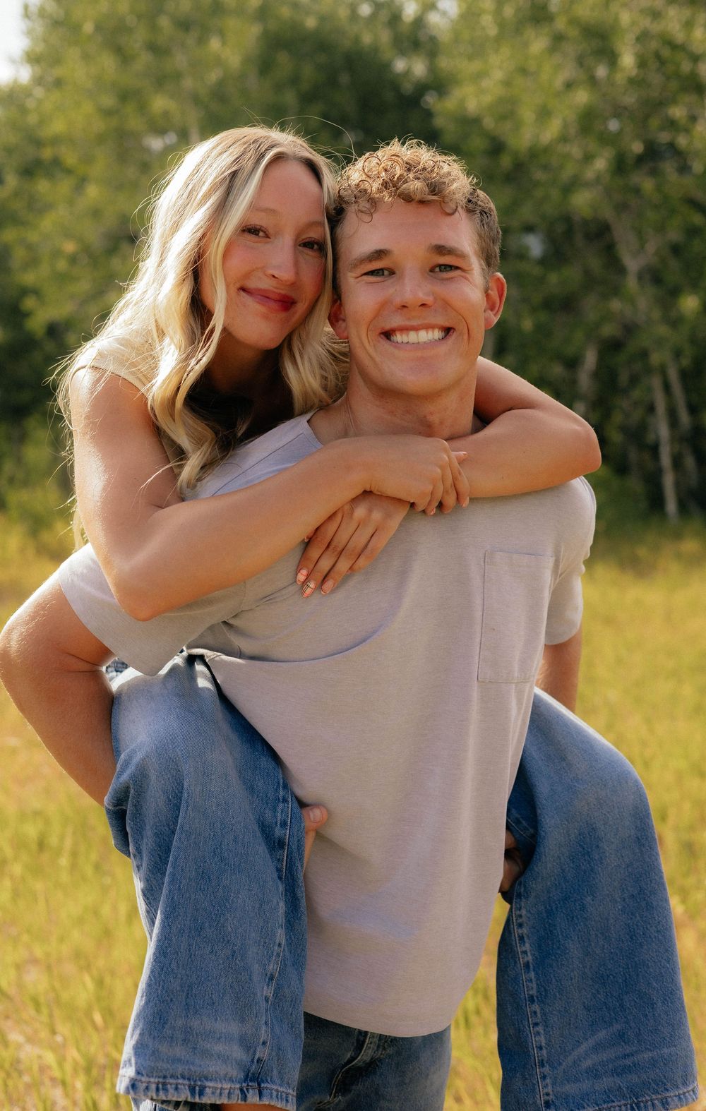 Happy couple posing in sunny green field, woman riding piggyback on smiling man in gray t-shirt and jeans.