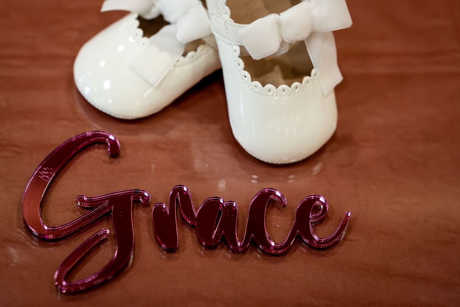White patent leather baby shoes with bows beside a shiny red cursive Grace name sign on brown surface.