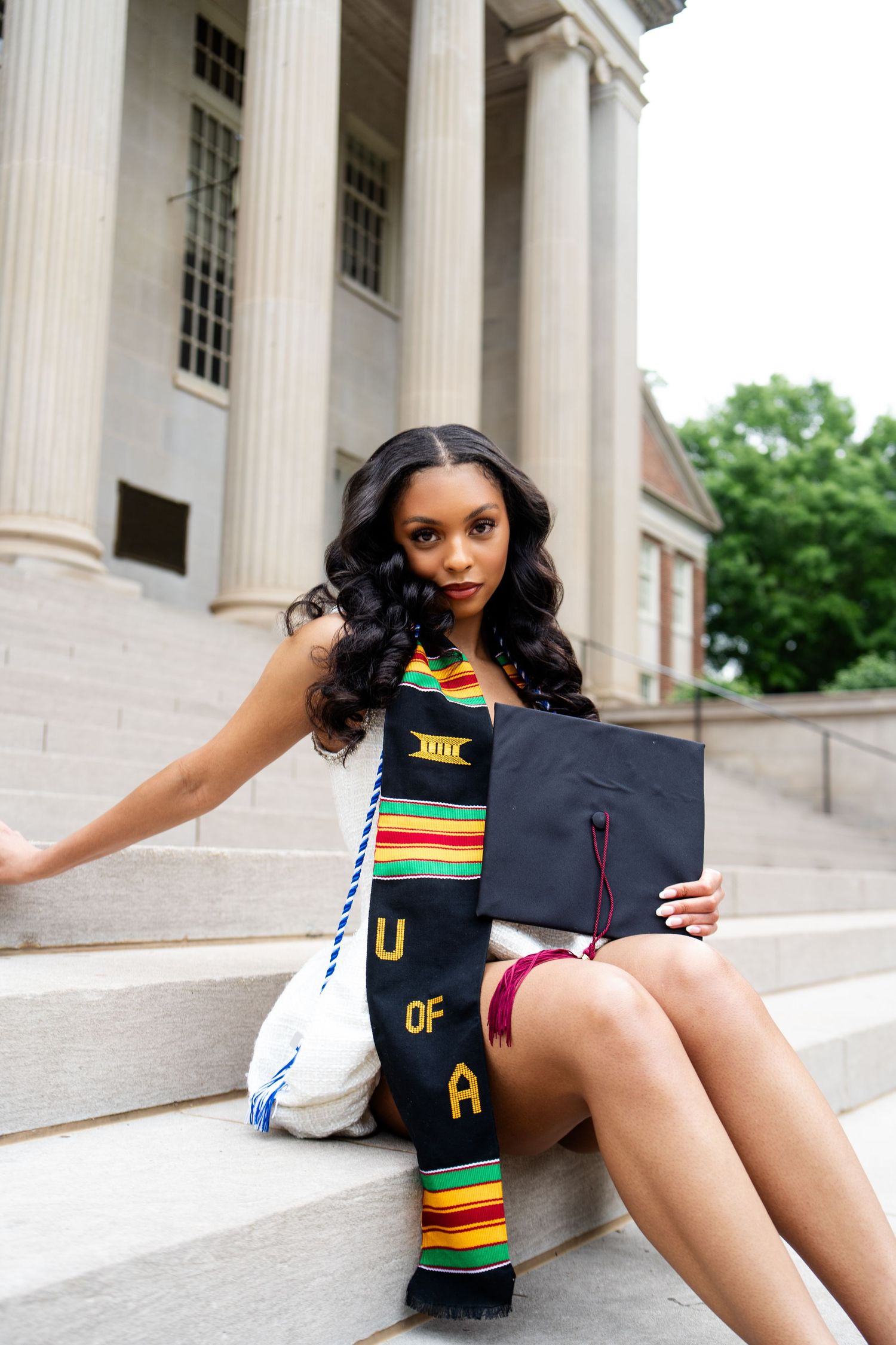 Smiling graduate in red dress with tattoos sits on stone steps holding black cap with red tassel.
