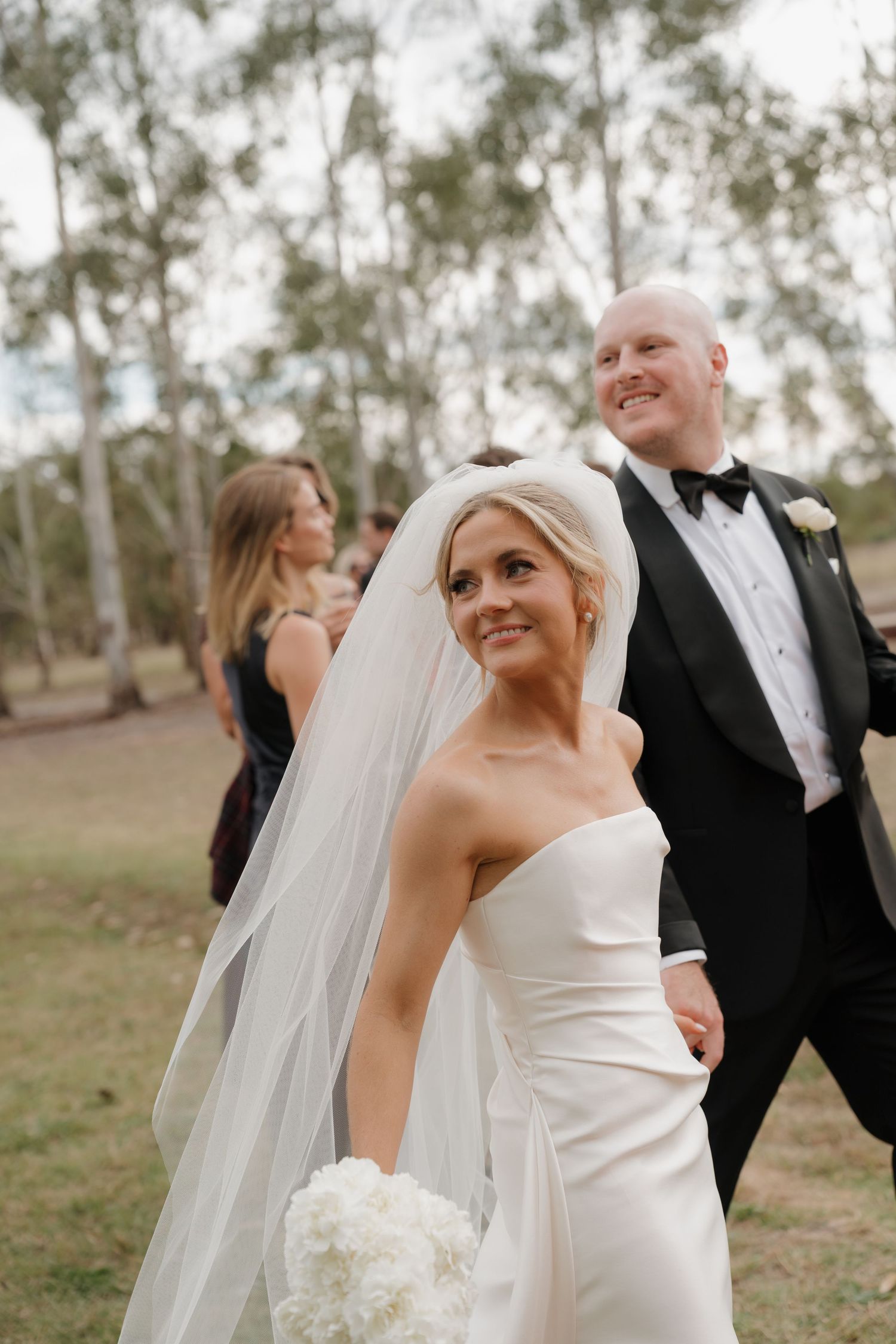 Bride in strapless white gown and veil holds bouquet while smiling, groom in black tuxedo beside her outdoors.