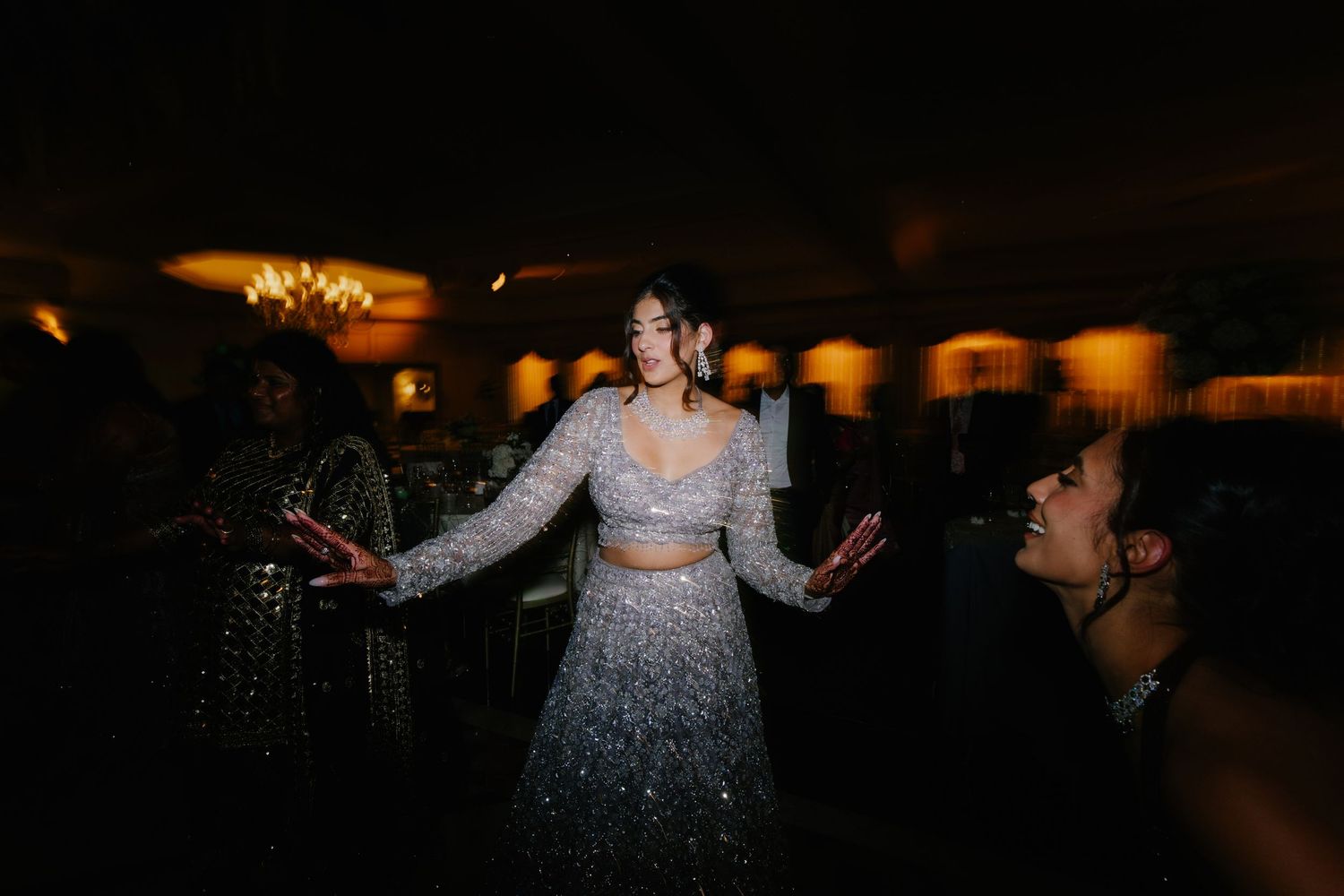 Black and white photo of a woman in embellished lehenga dancing joyfully surrounded by guests at an Indian wedding celebration.