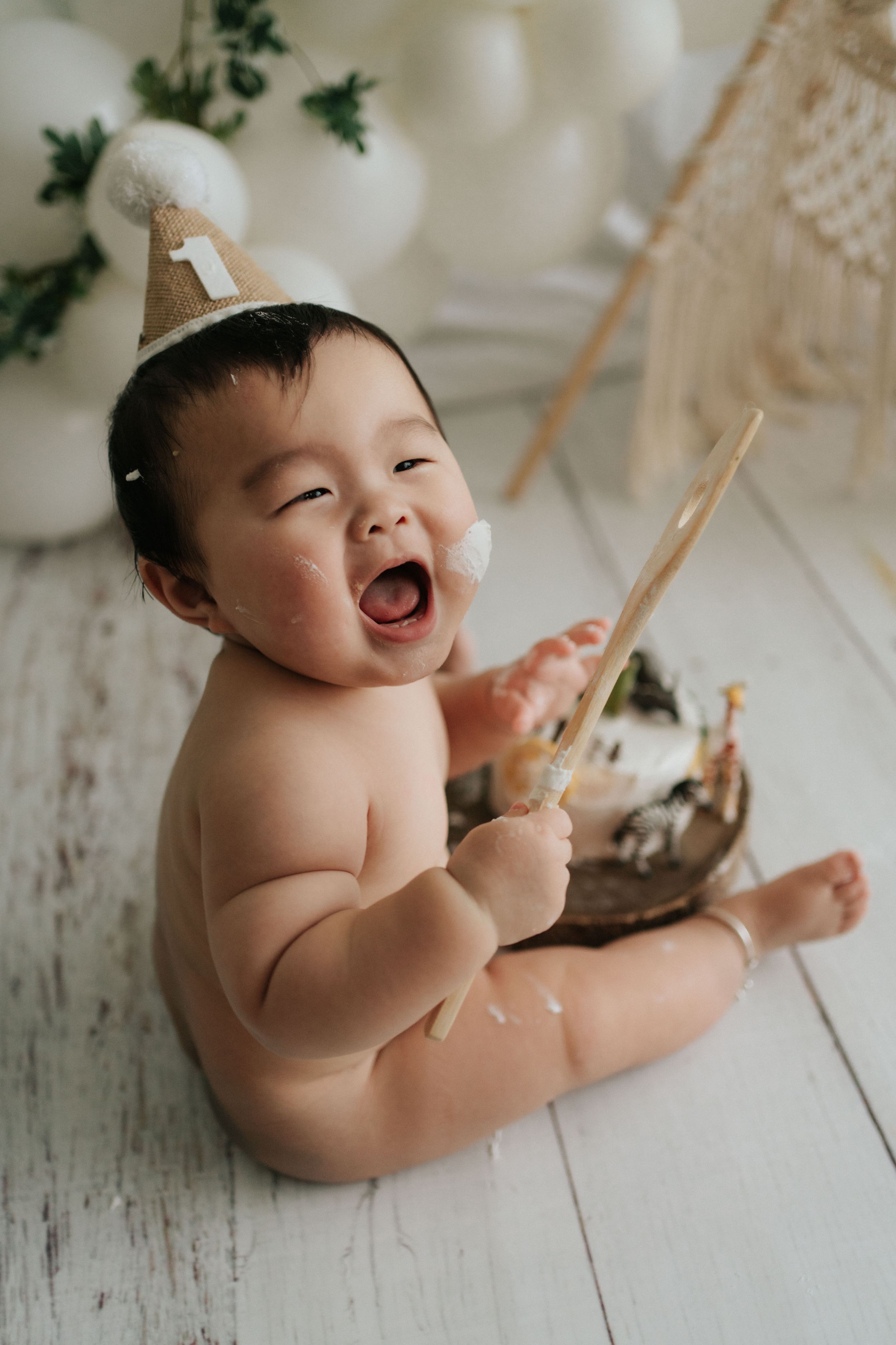Baby wearing party hat sits on white floor with excited expression.戴著派對帽的嬰兒興奮地坐在白色地板上。