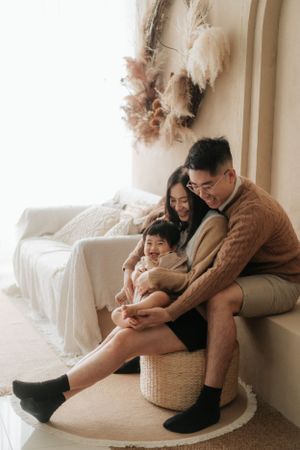 A happy Asian family of three sits together on a wicker stool, smiling at a phone in a cozy, neutral-toned room.一個幸福的亞洲三口之家坐在藤凳上，在一個溫馨的、色調柔和