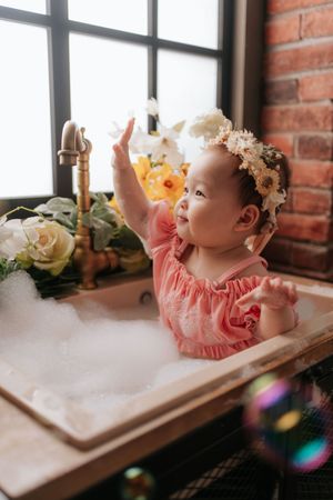 Adorable baby girl in pink dress with floral crown sits in bubble bath reaching toward brass faucet surrounded by flowers.可愛的女嬰穿著粉紅色連身裙，頭戴花冠