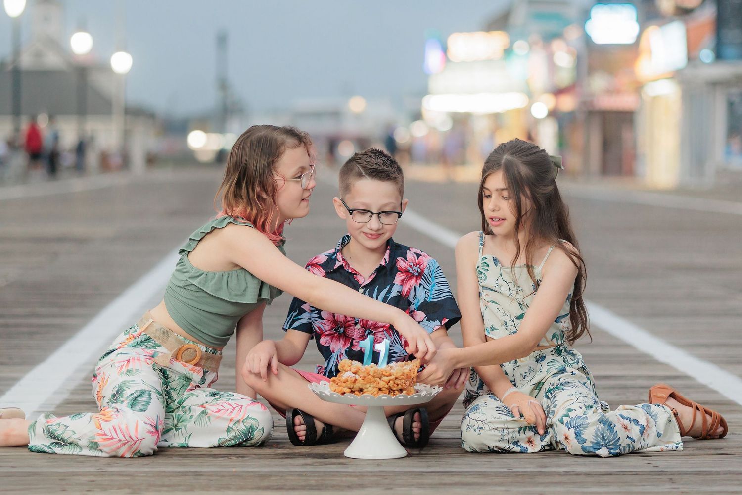 Three children sit on a boardwalk celebrating a birthday with a small cake and candles at dusk.