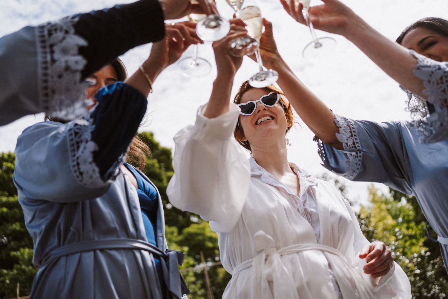 Bride and bridesmaids in robes toasting with champagne glasses outdoors at a wedding celebration.