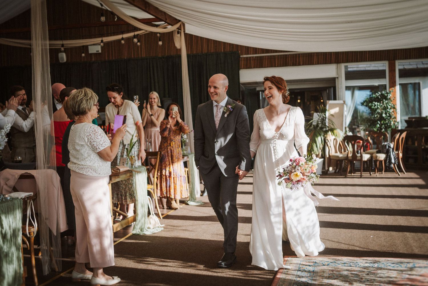 Bride and groom walk down decorated aisle as smiling guests celebrate at elegant outdoor wedding.