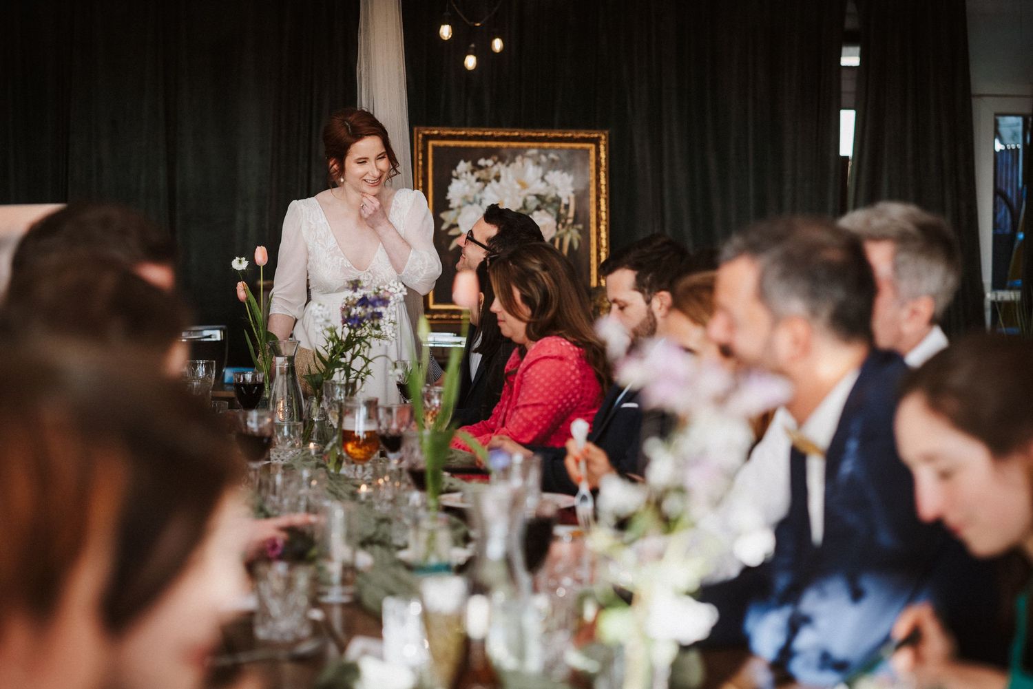 Bride laughs while giving a wedding speech to elegantly dressed guests seated at a long floral-decorated reception table.