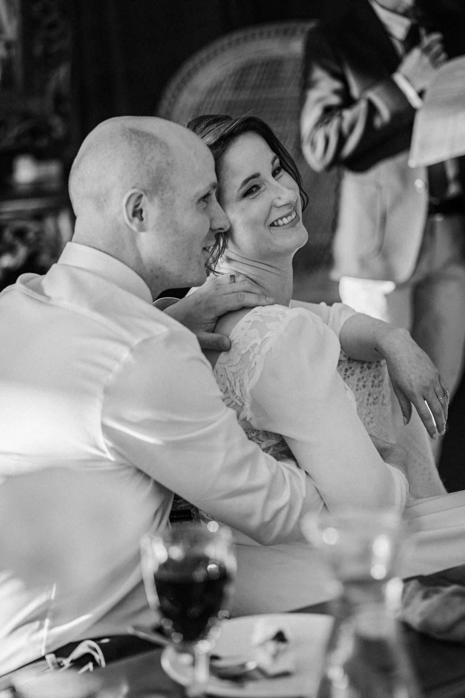 Black and white photo of groom embracing laughing bride at wedding reception table.