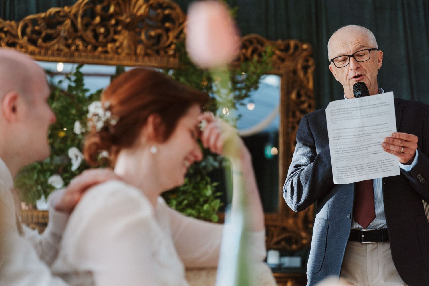 Elderly man reads speech at wedding reception while bride and groom listen at decorated table.