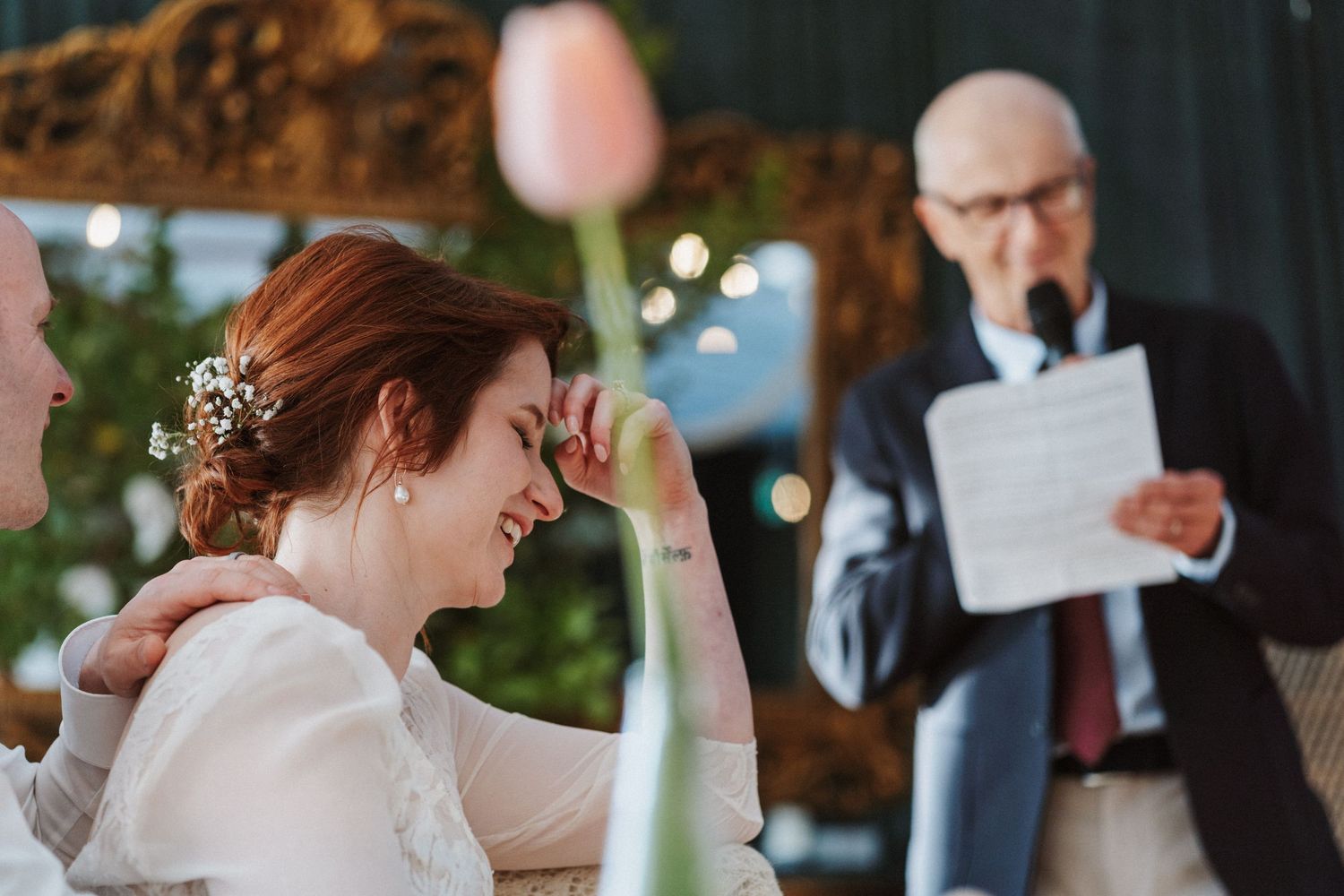 Emotional bride laughs during wedding speech while seated next to groom at reception table.