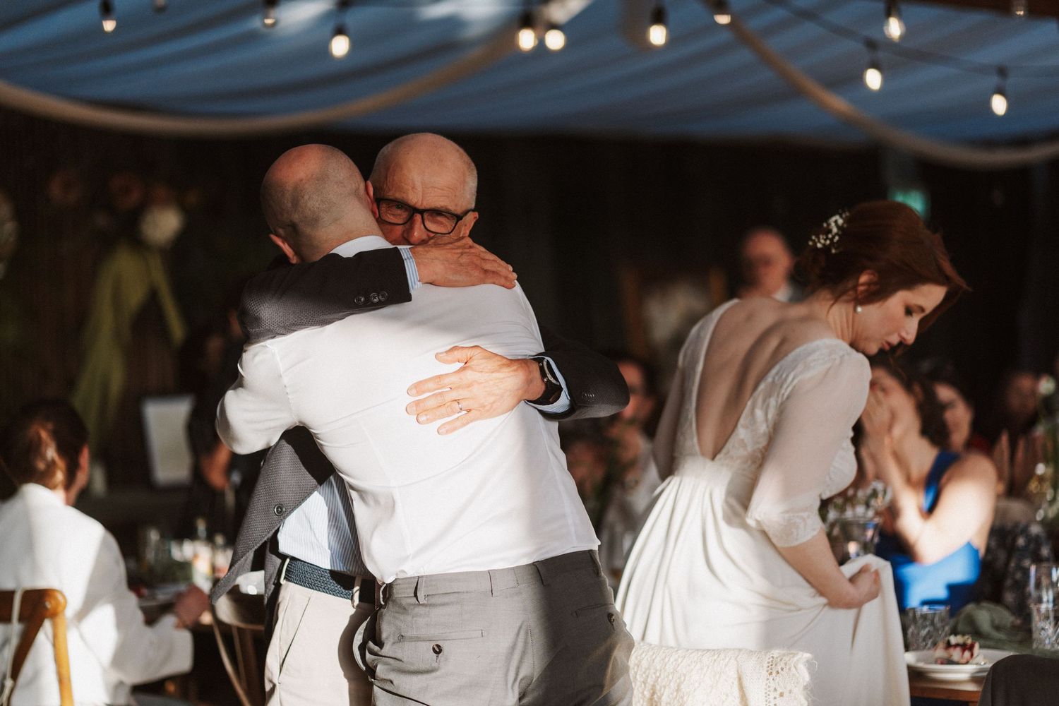 Bald groom in white shirt shares emotional hug with guest while bride in lace dress stands nearby.