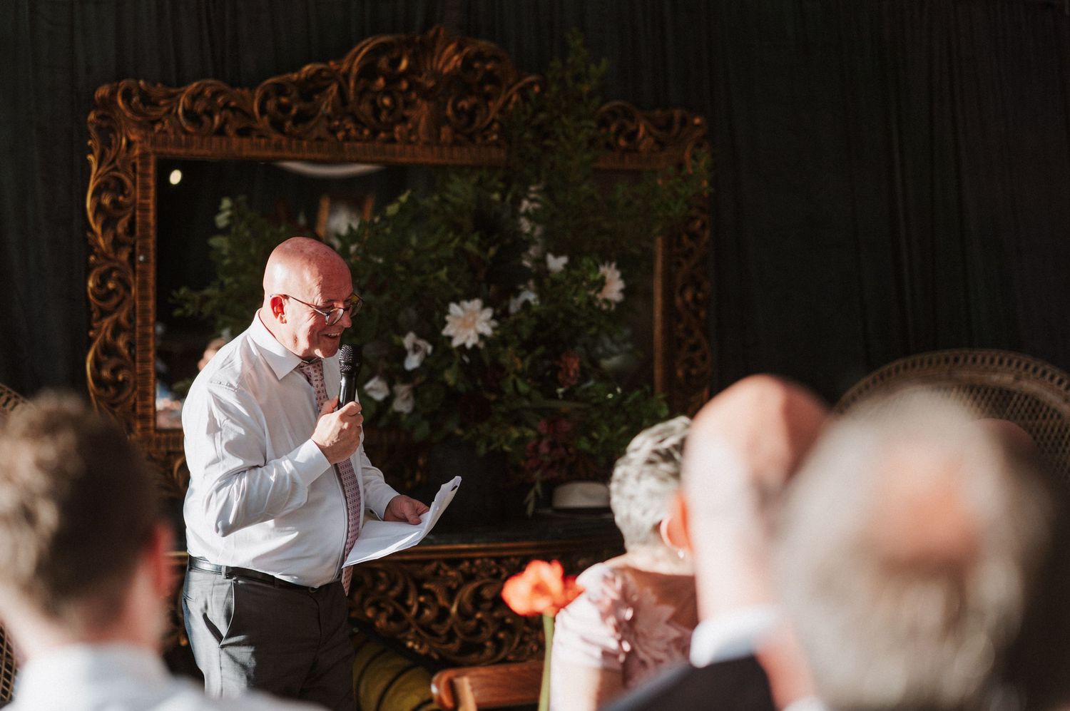 Bald man delivers heartfelt wedding speech at microphone before ornate gold mirror with floral decor.
