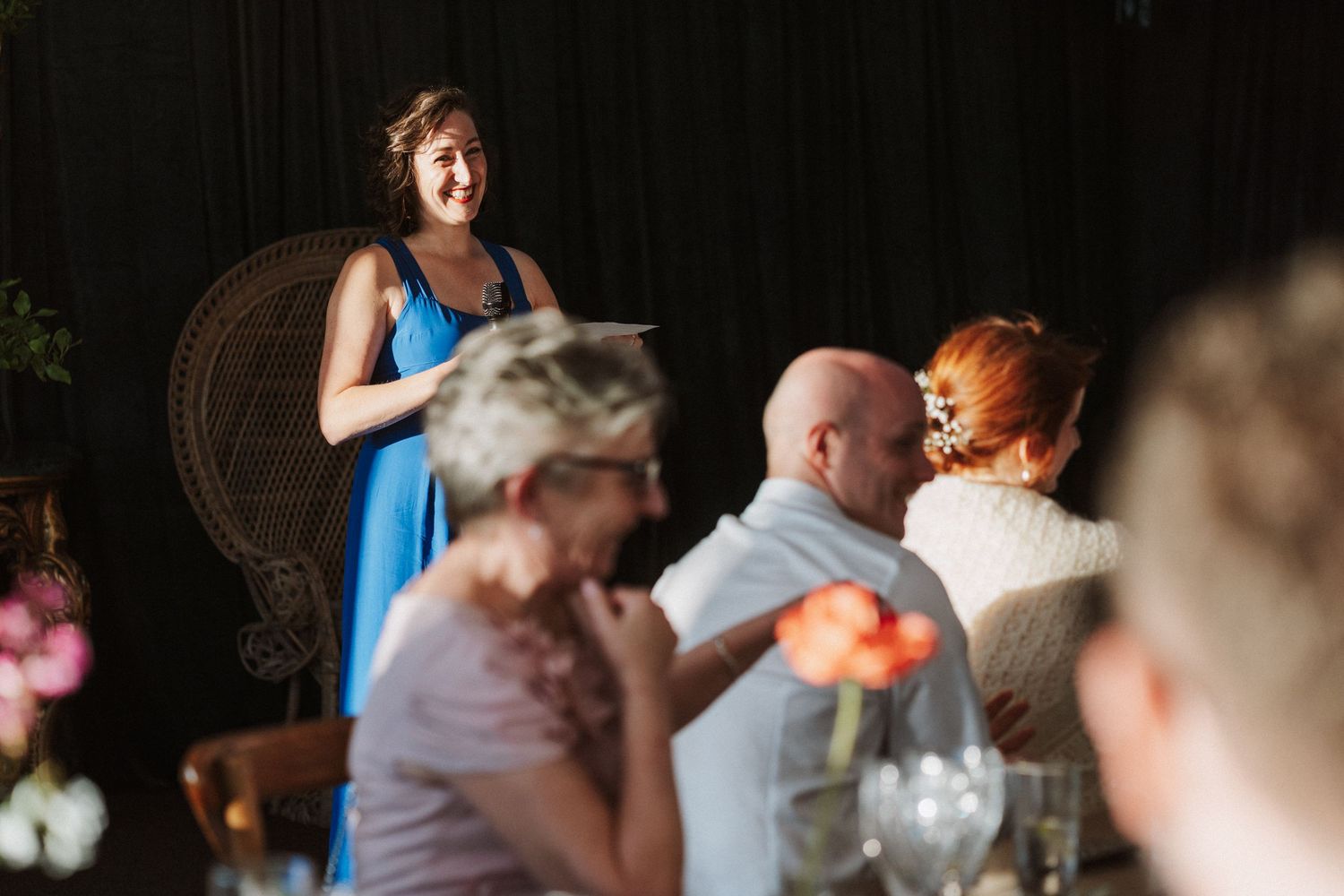 Woman in blue dress gives toast at wedding reception while guests smile and embrace at table.
