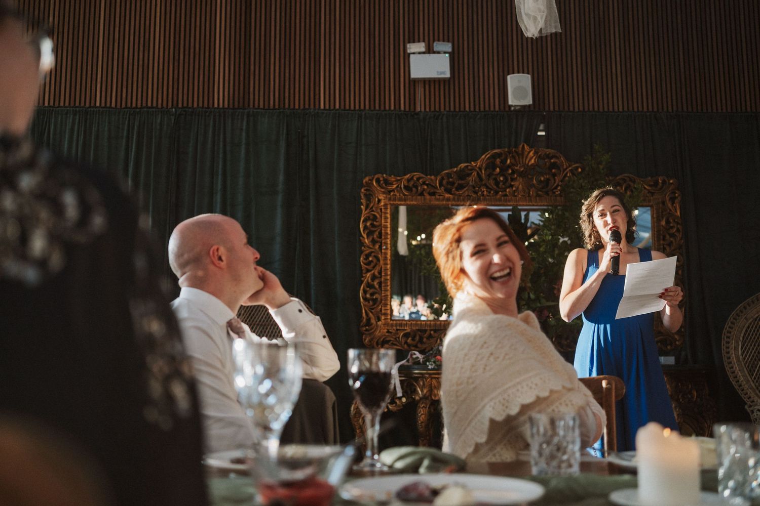 Bride laughs joyfully during wedding speech while groom smiles at decorated reception dinner.