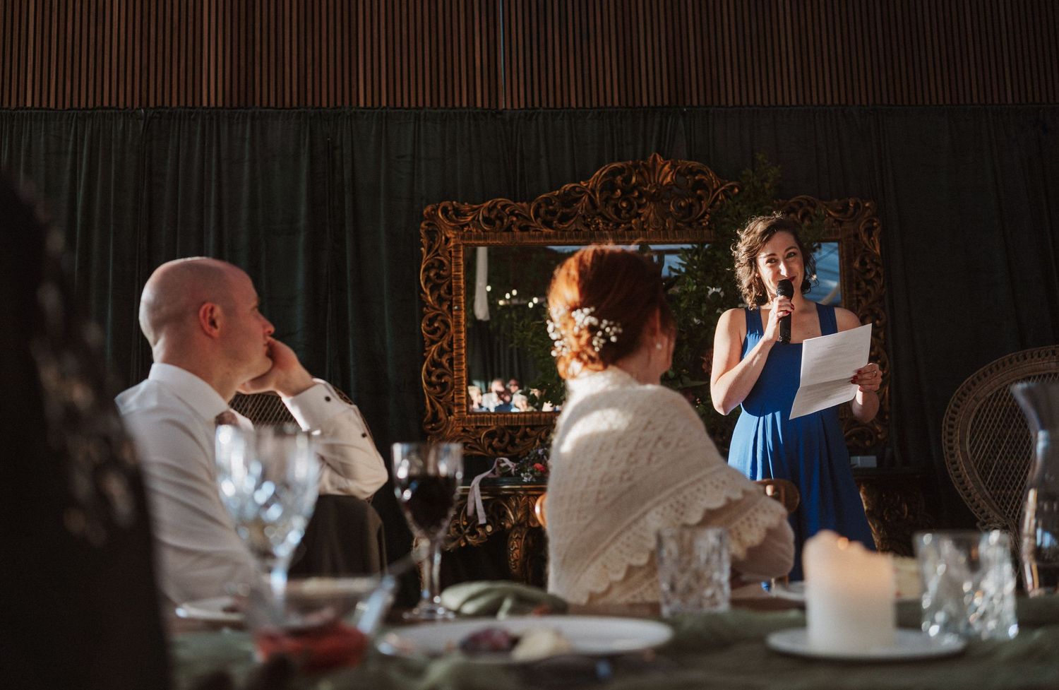 Wedding guest in blue dress gives a heartfelt speech at an elegant candlelit reception dinner.