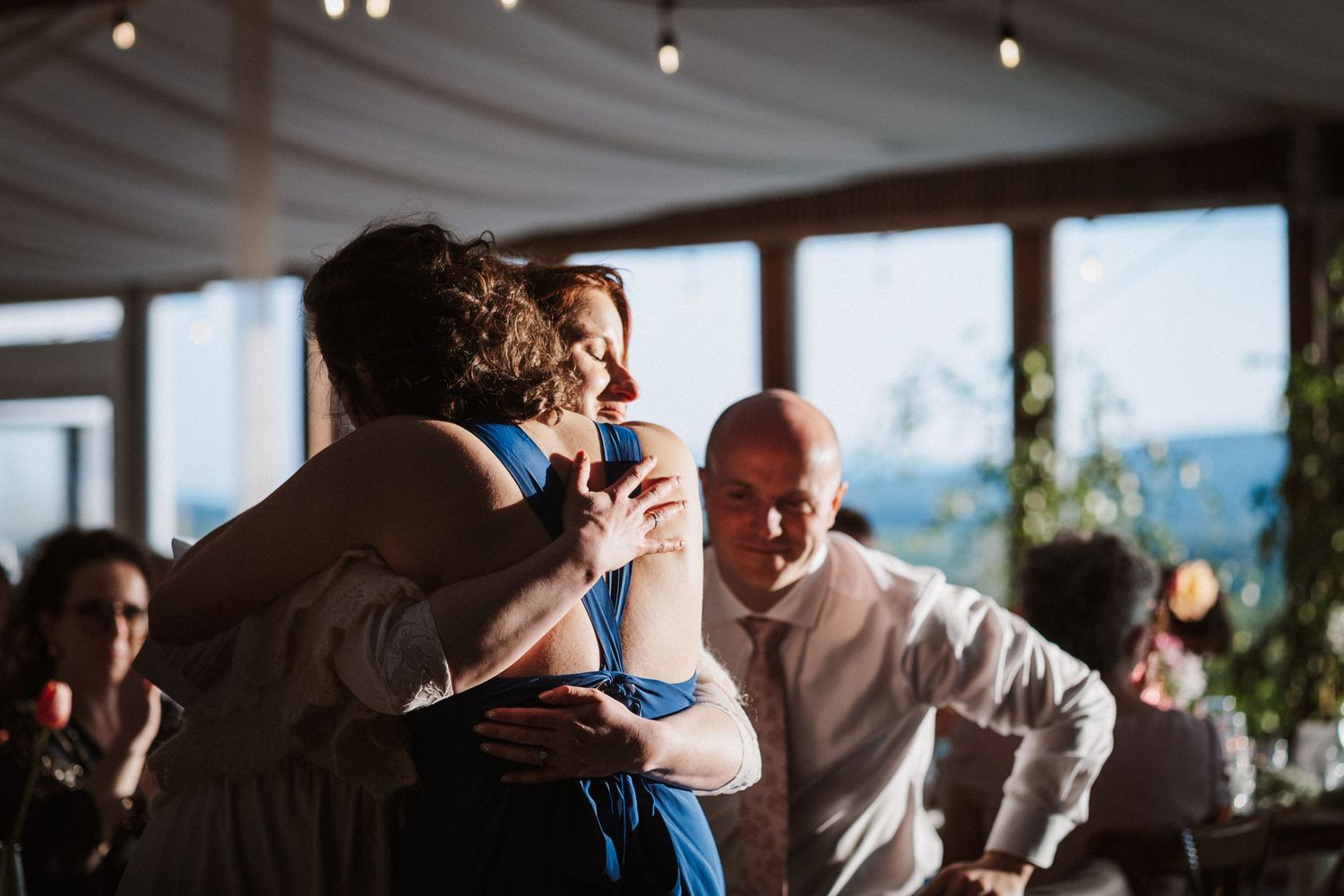 Wedding guests in blue dress embrace and dance together at lively indoor reception party.