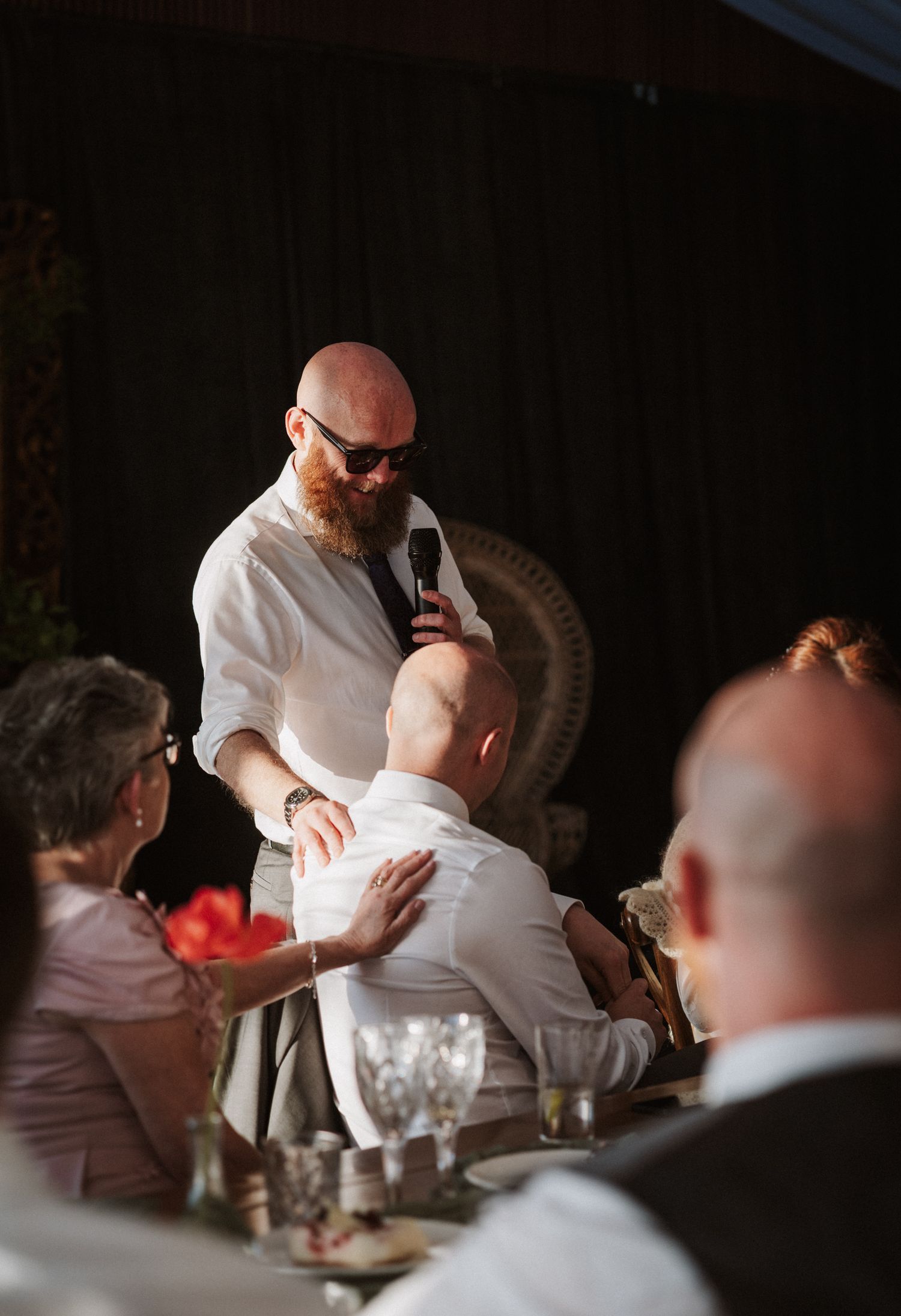 Bald bearded man with sunglasses gives speech at wedding holding microphone over seated guests.