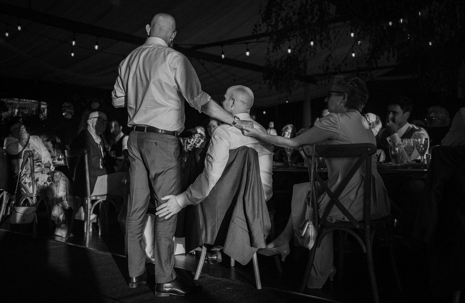 Black and white photo of wedding guests dancing energetically under string lights at a reception.