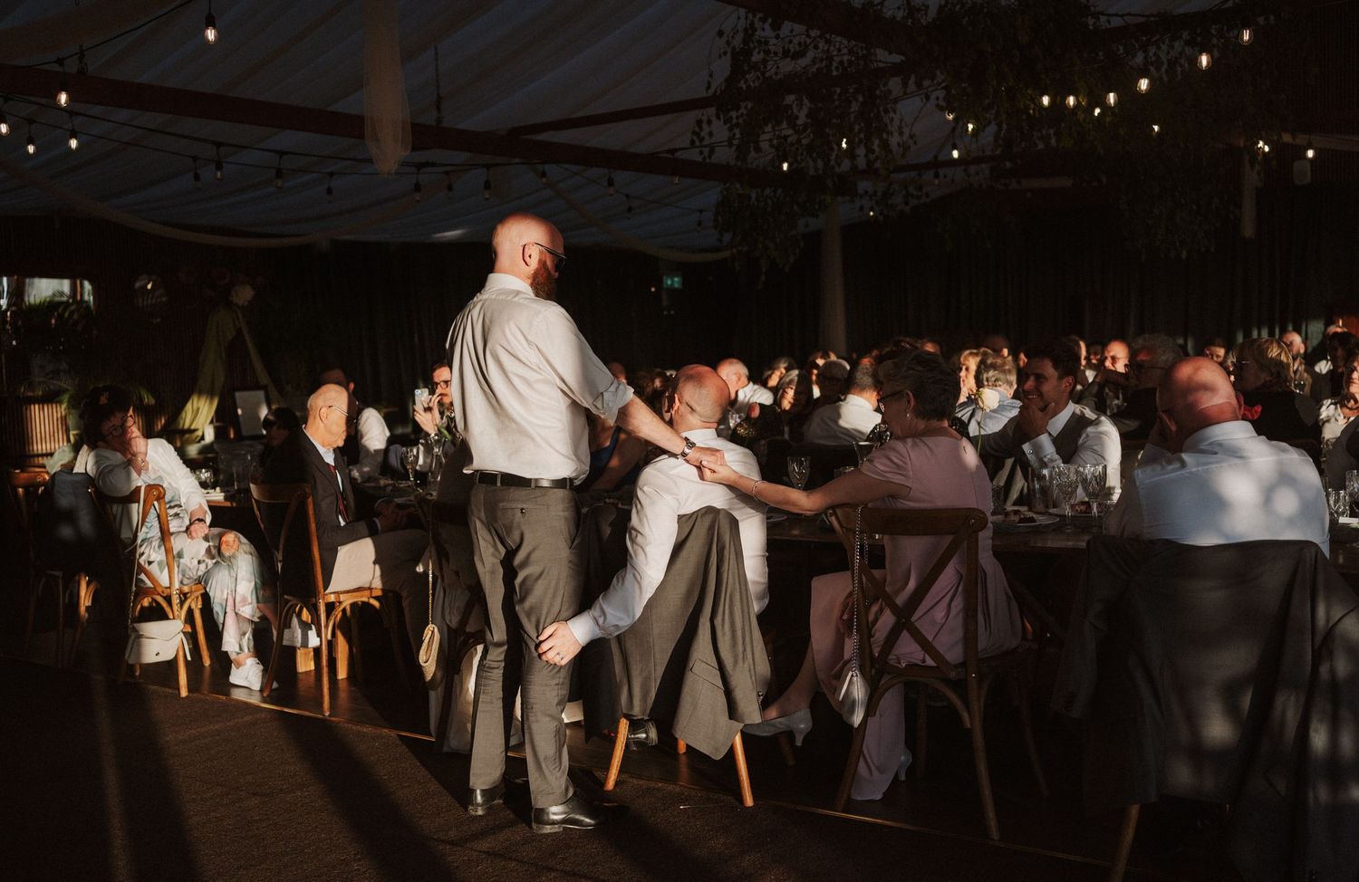Wedding guests dance and celebrate under warm string lights inside a large tent reception venue.