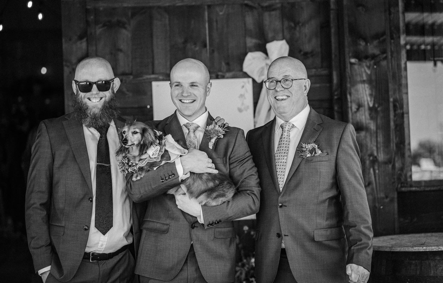 Three men in suits smile for a wedding photo, with the groom holding an adorable dog in the center.