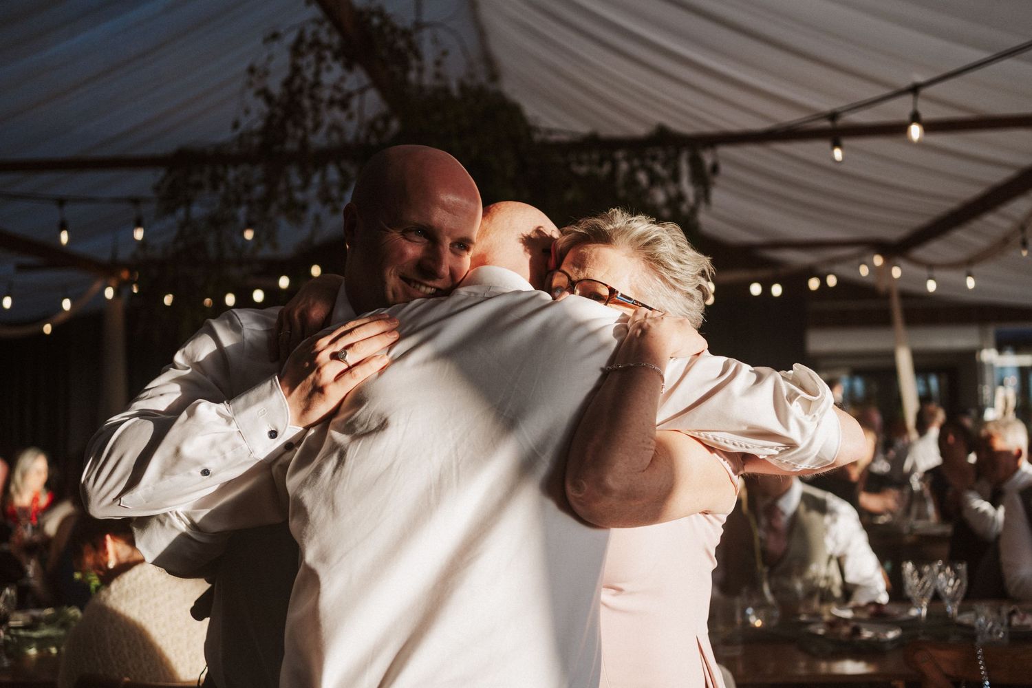Emotional group hug between bride, groom and guest under twinkling lights at wedding.