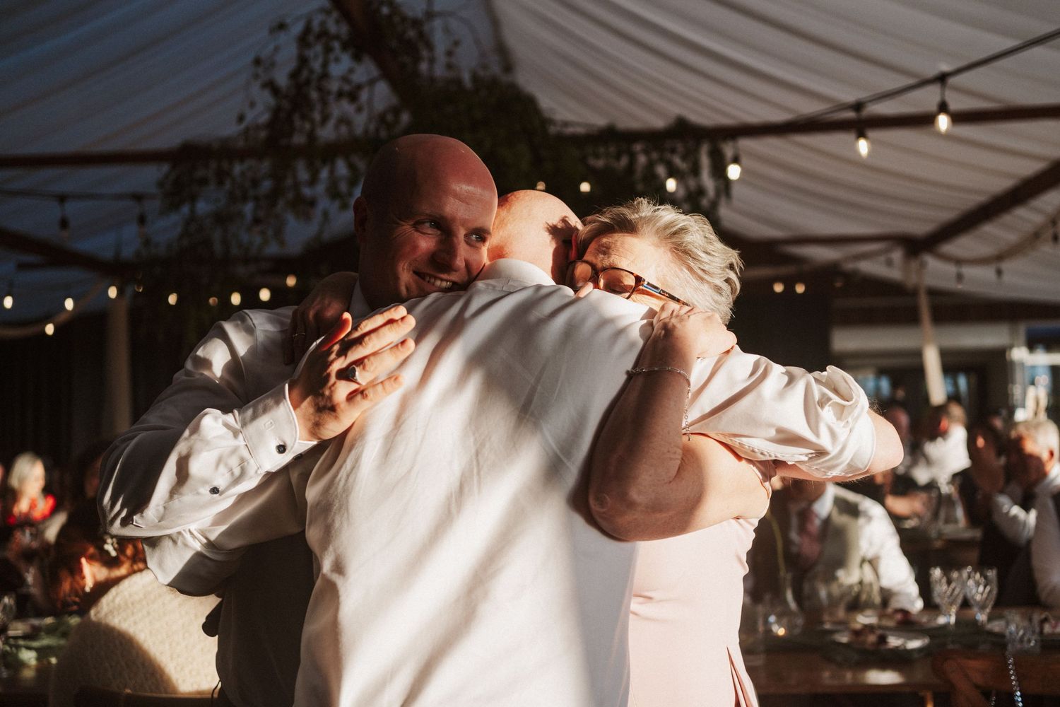 Bald groom shares a warm group hug with two guests under string lights at a wedding reception.
