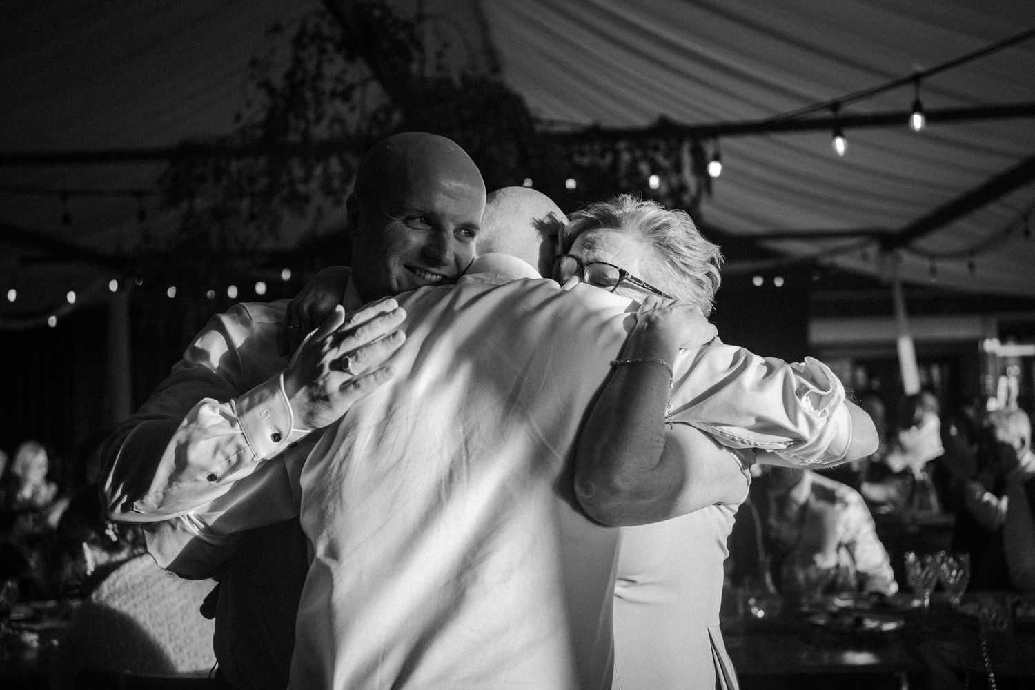 Black and white photo of joyful group hug between three people at outdoor wedding reception.