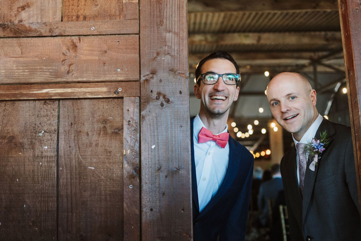 Two grooms smiling joyfully in a rustic barn doorway, one wearing a pink bow tie and glasses at a wedding celebration.