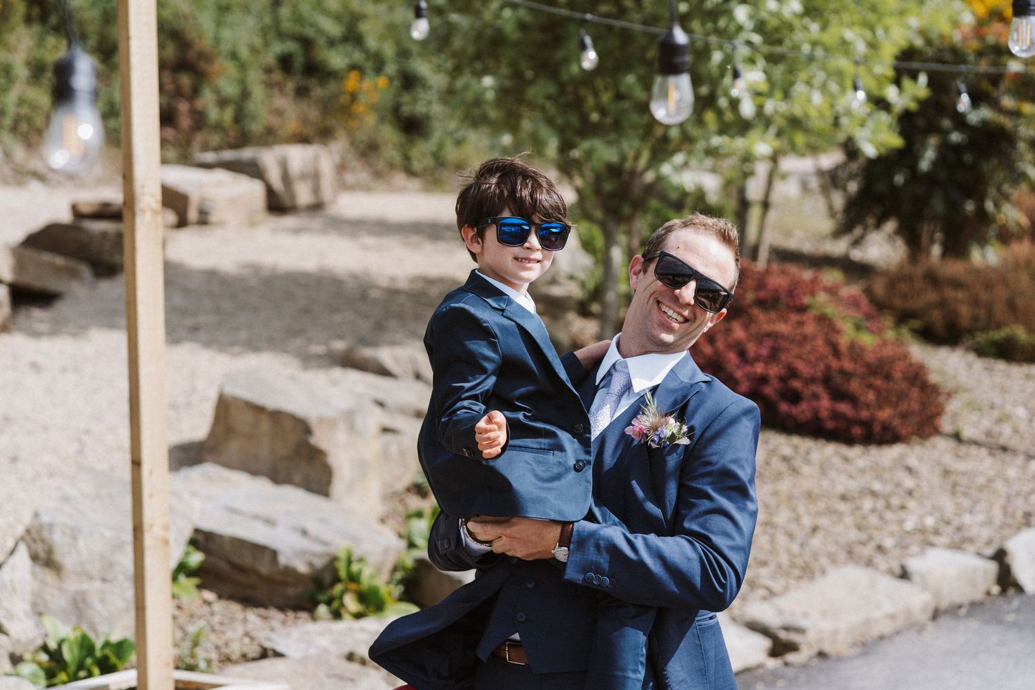 Groom in blue suit holds young boy in matching suit, both wearing sunglasses at outdoor wedding venue.
