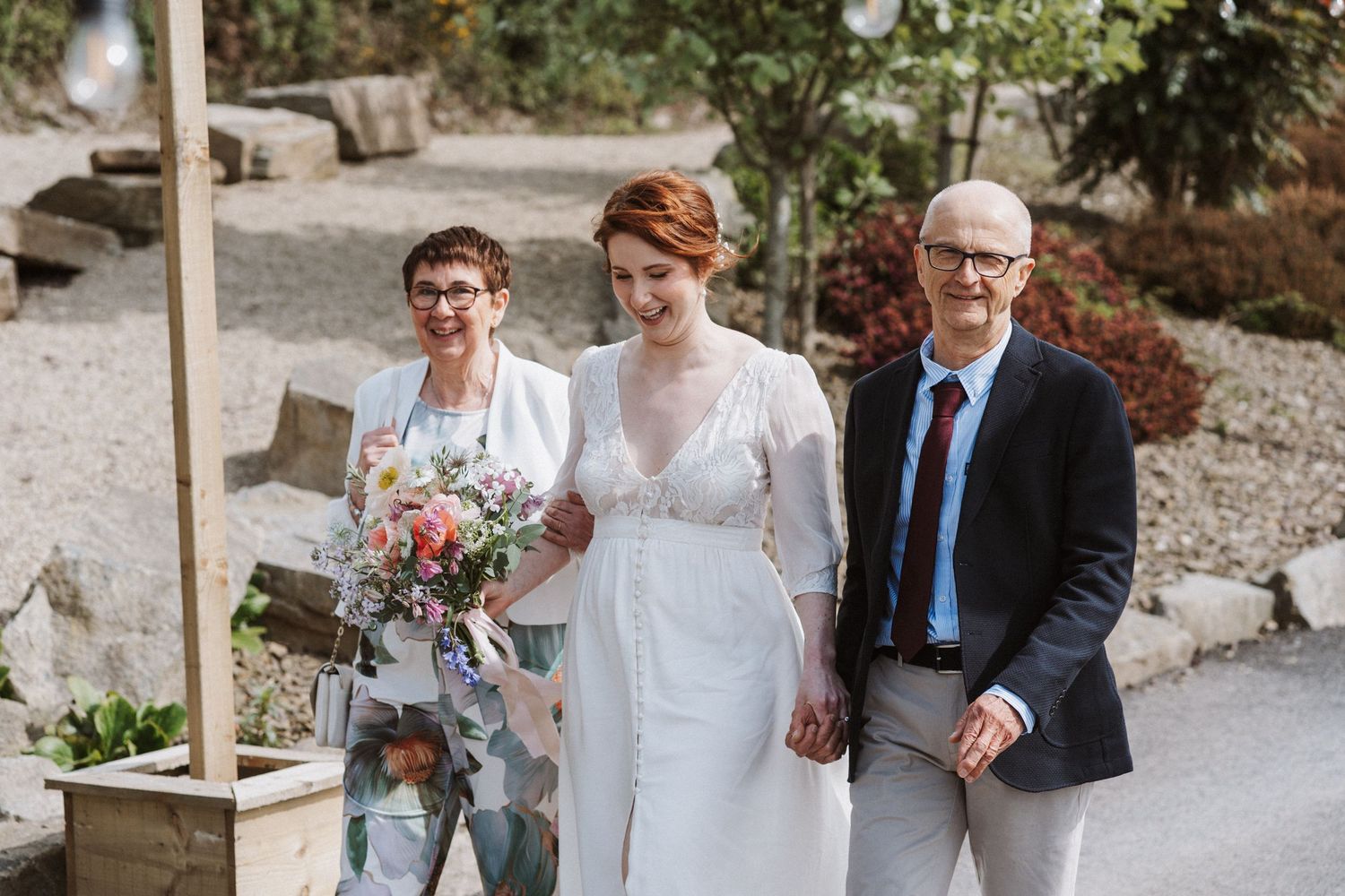 Bride in white gown walks with parents toward wedding ceremony outdoors, holding colorful bouquet.