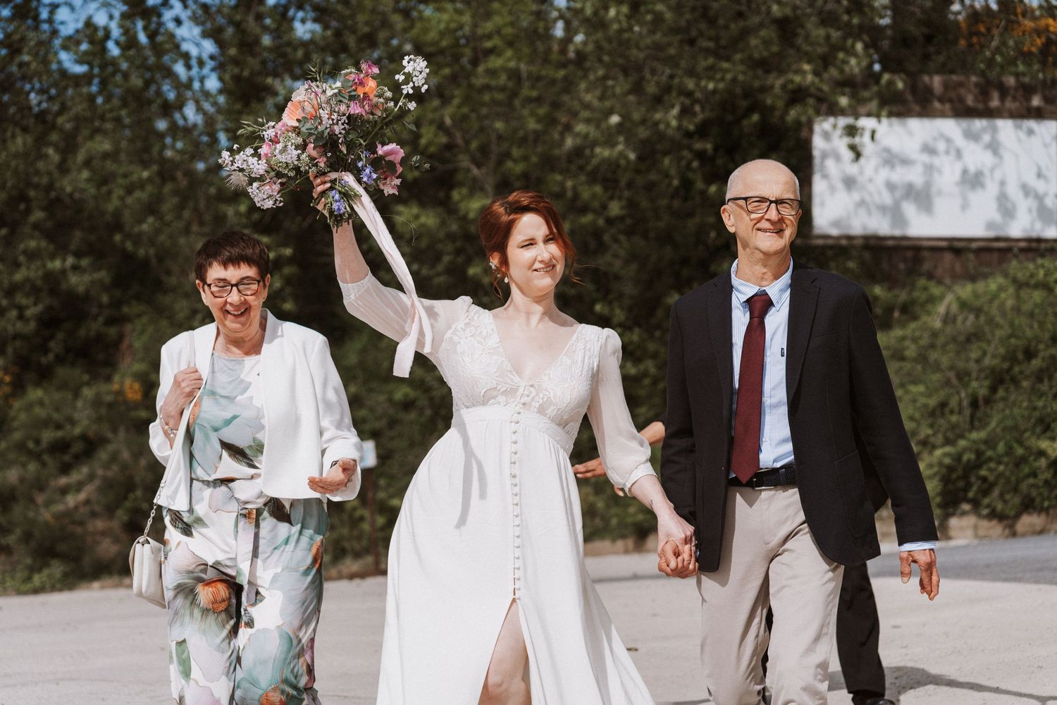 Bride in white gown raises colorful bouquet while walking with parents at outdoor wedding celebration.