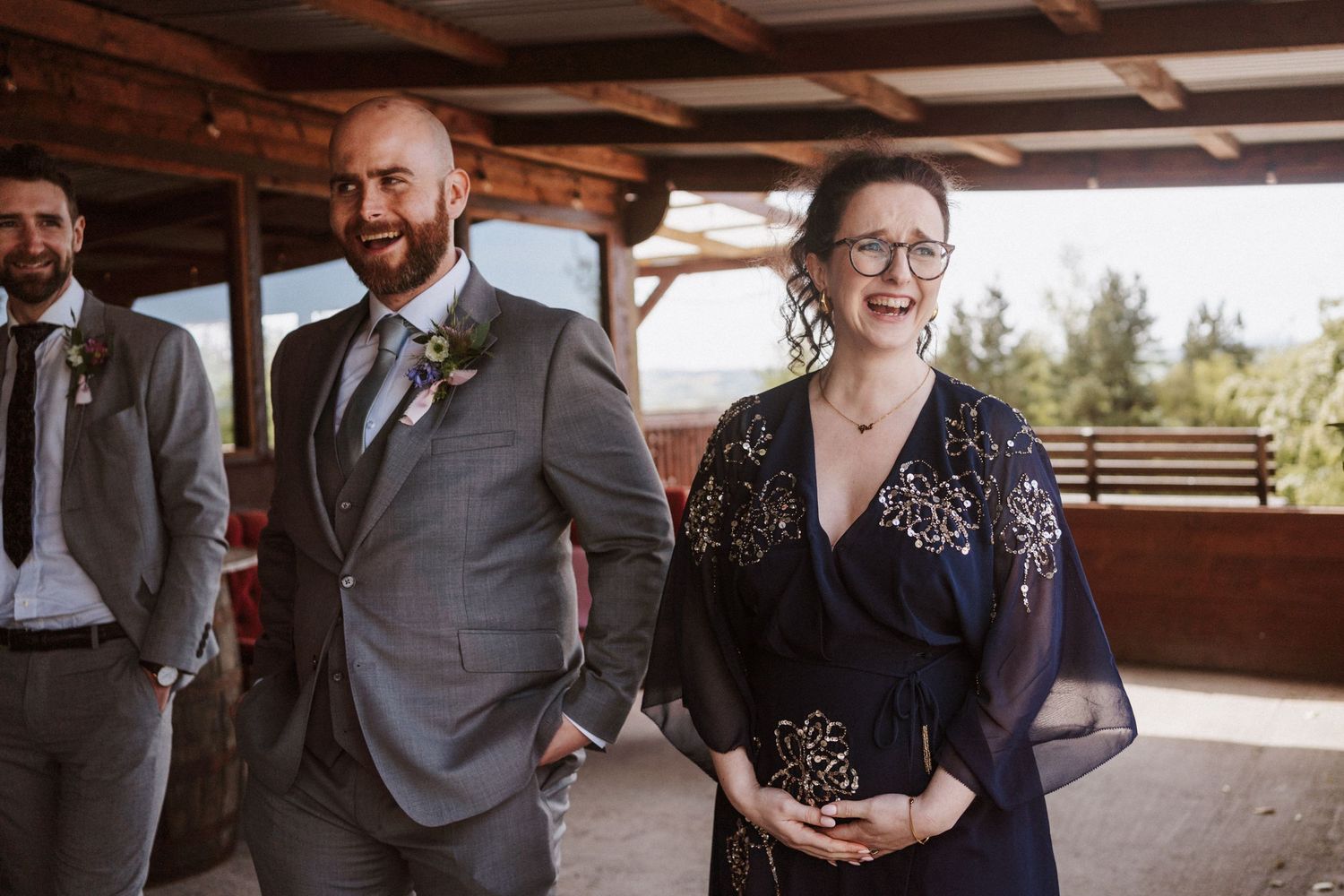 Groom in gray suit smiles with woman in navy sequin dress at outdoor wedding venue.