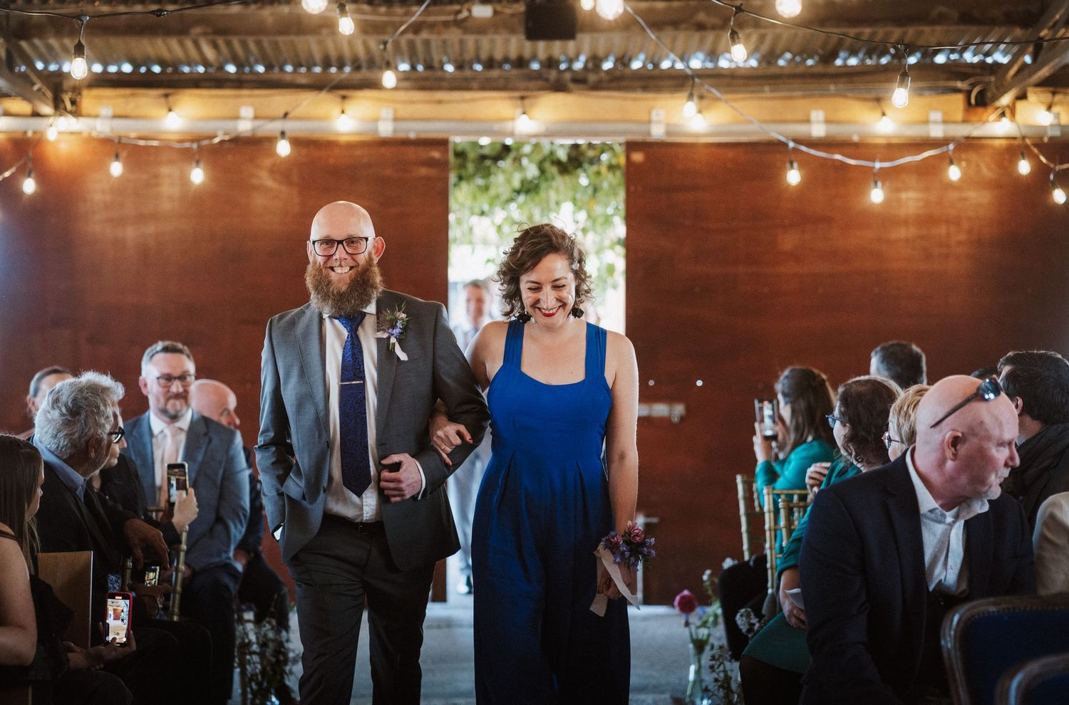 Couple walks arm-in-arm at an indoor wedding reception with string lights and wooden walls.