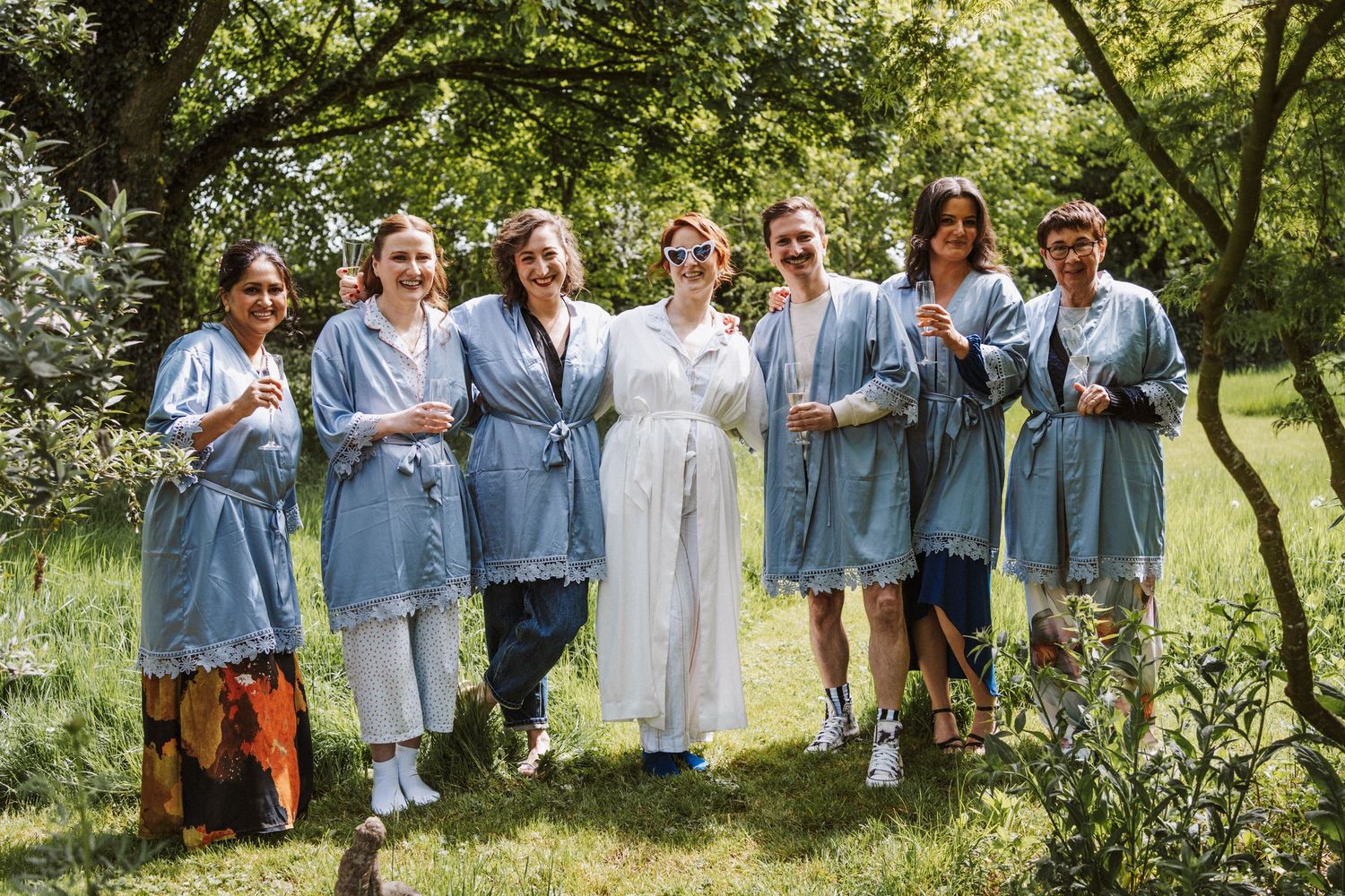 Bride and bridesmaids in matching blue lace robes smiling outdoors in a lush green garden setting.