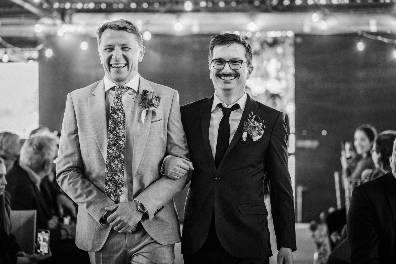 Two grooms smiling joyfully in suits with boutonnieres at their wedding ceremony in black and white photo.