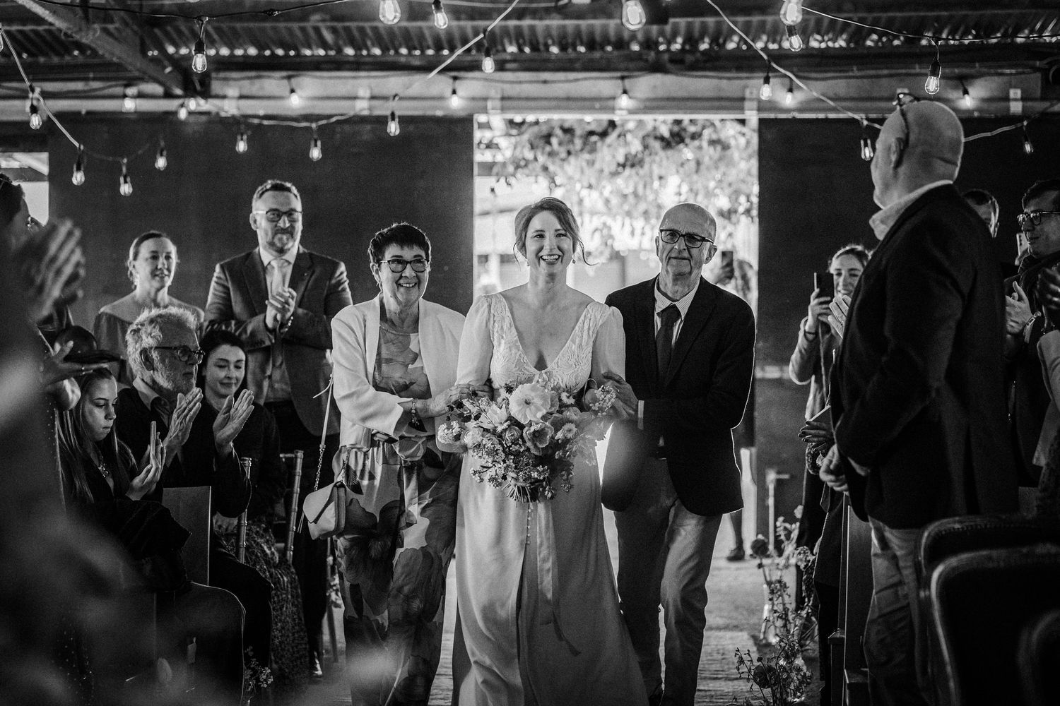 Black and white photo of a bride walking down the aisle with family, guests applauding in a warmly lit venue.