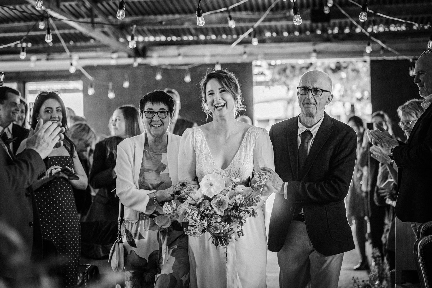 Bride walks down the aisle with parents in black and white wedding ceremony photo.