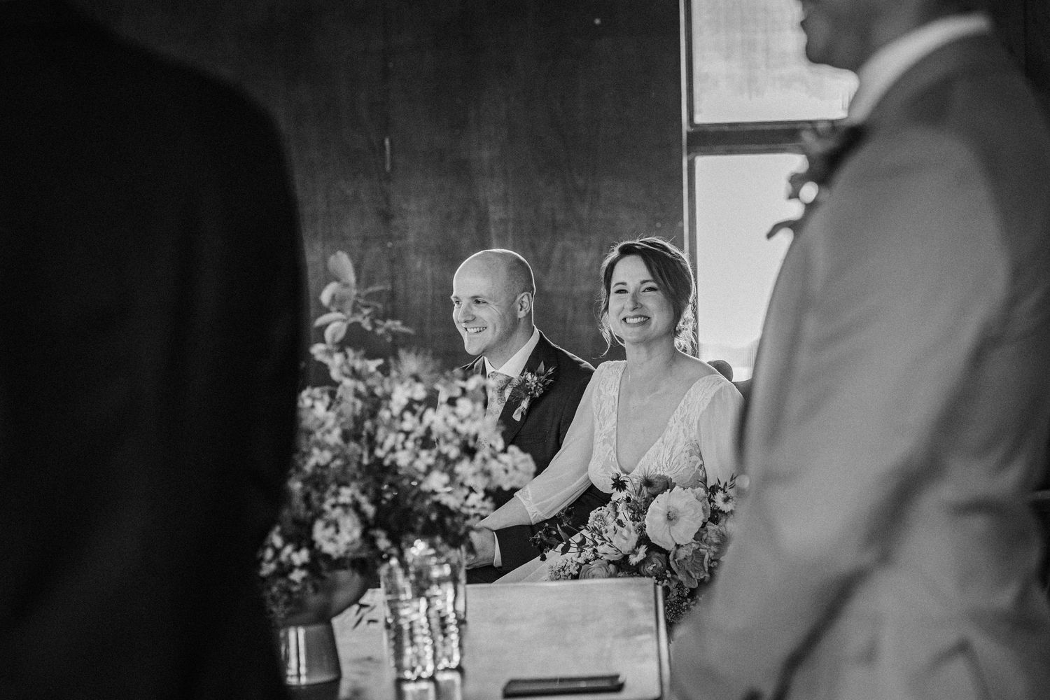 Black and white photo of a smiling bride and groom during their wedding ceremony, seated together at a table.