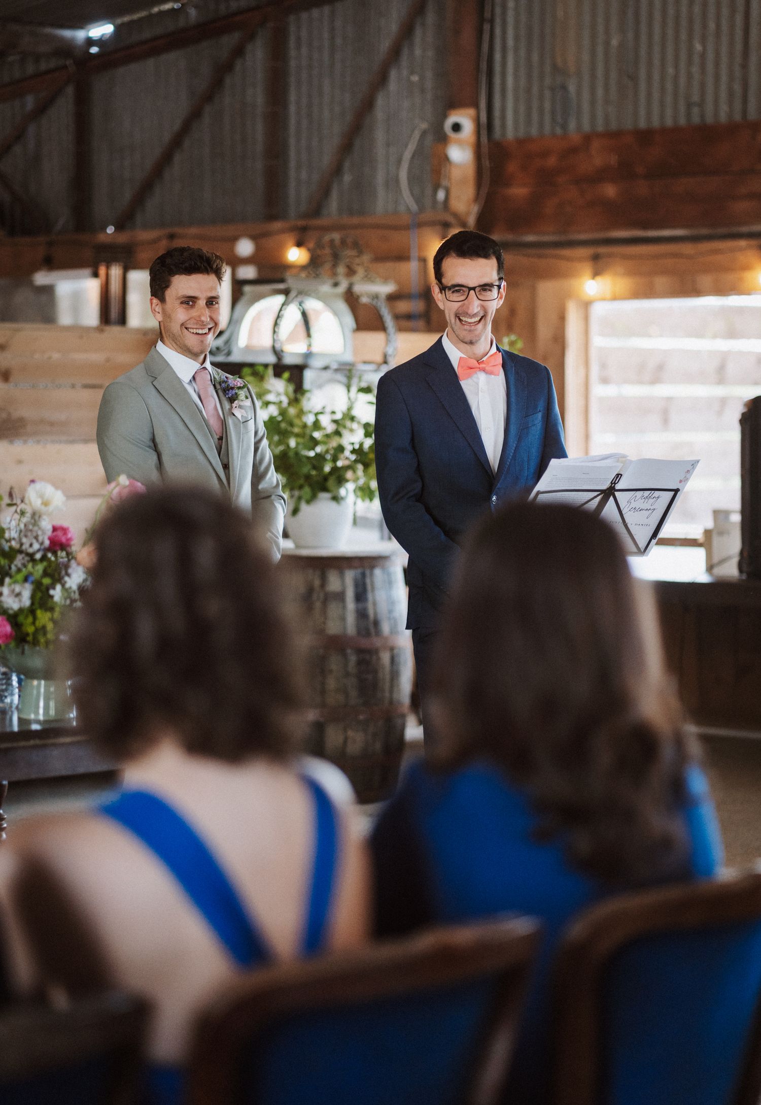 Two grooms smiling during their barn wedding ceremony while guests watch from decorated wooden chairs.