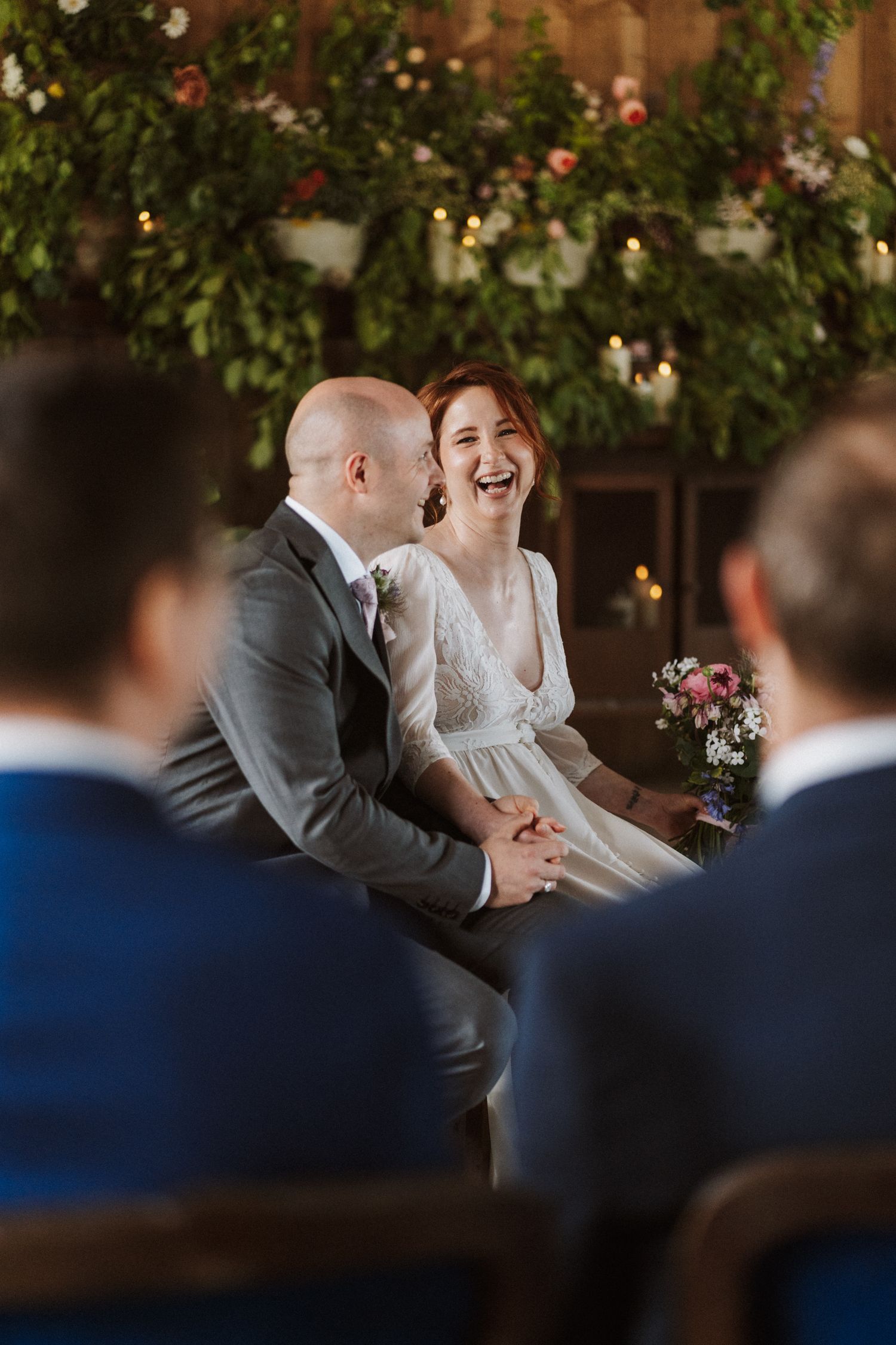 Bride laughing joyfully with groom during intimate wedding ceremony, surrounded by lush greenery and floral decorations.