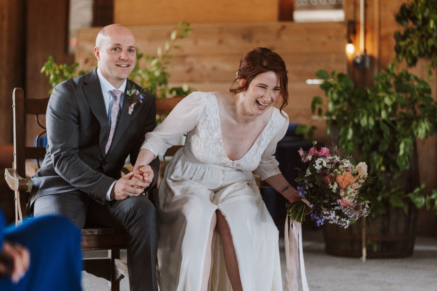 Bride and groom laughing joyfully while holding hands seated at rustic barn wedding ceremony.